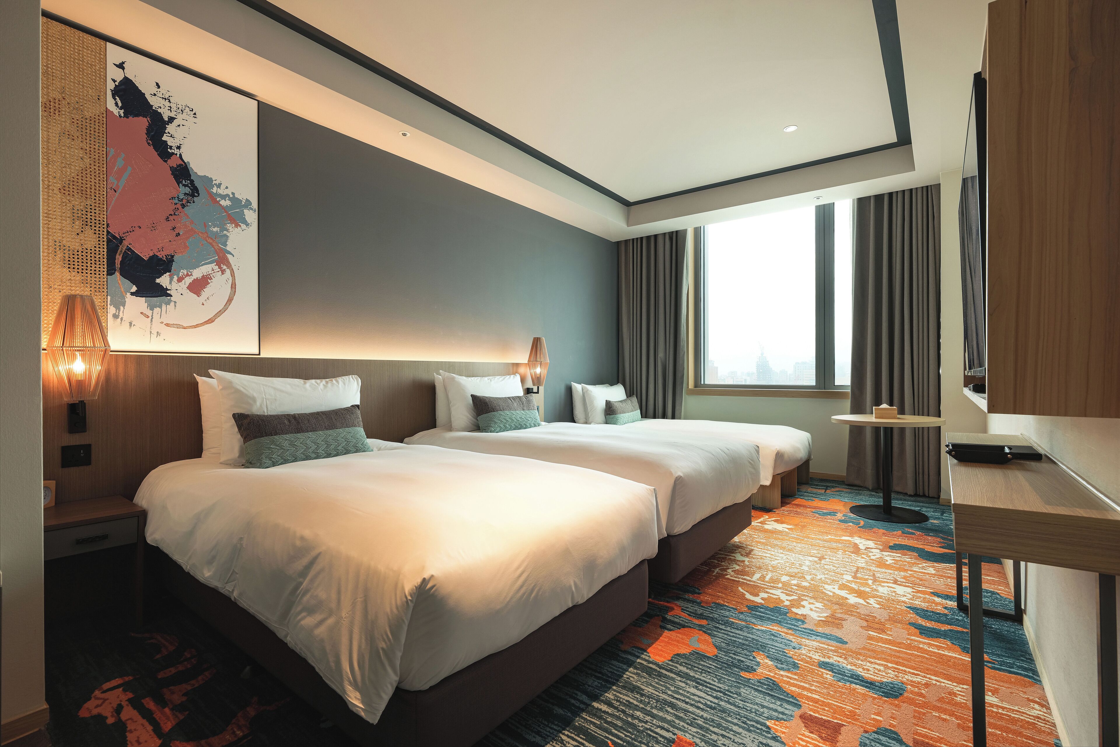 A hotel room with two beds, a large abstract painting, a desk, and a view of the city through the window.