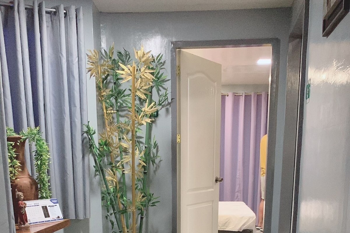 Un pasillo con suelo pulido, un escritorio con computadora, una planta en maceta y una cama al fondo.