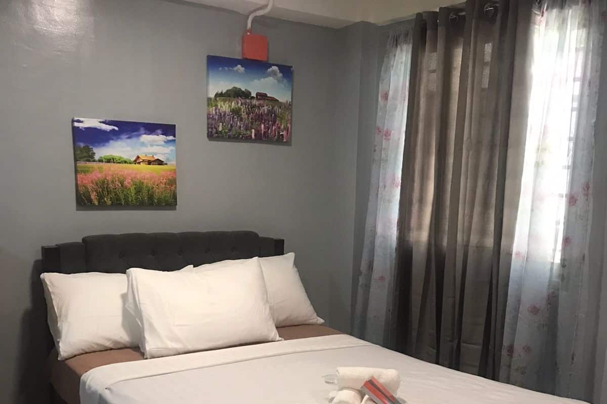 Una cama bien tendida con ropa blanca y un cabecero marrón, una alfombra marrón al pie de la cama y dos cuadros enmarcados en la pared.
