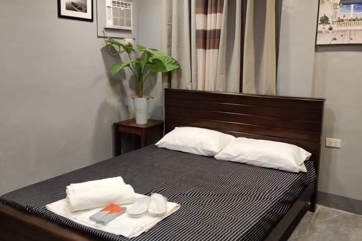 Una cama bien tendida con una colcha a rayas, una planta en la mesita de noche y una pequeña mesa con una lámpara.