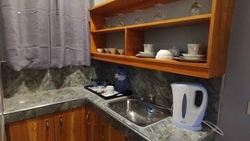 A compact kitchen with wooden cabinets, a marble countertop, a built-in shelf with glassware, and a kettle.