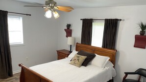 Un lit bien fait, avec une tête de lit en bois, une table de chevet avec une lampe, un ventilateur de plafond et une fenêtre avec des rideaux.