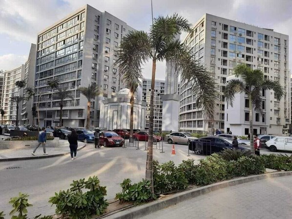 A modern apartment building with multiple floors, a parking area with cars, and palm trees.