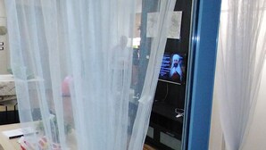 Una stanza con un'ampia finestra ornata da tende bianche trasparenti, un pavimento in legno e una televisione sullo sfondo.