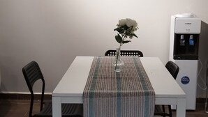 Zona pranzo con tavolo bianco, sedie nere, tovaglia fantasia, un vaso con fiori bianchi e un dispenser d'acqua.