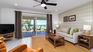 Una sala de estar moderna con sofá, mesa de centro y televisor, un balcón con mobiliario exterior y un ventilador de techo.