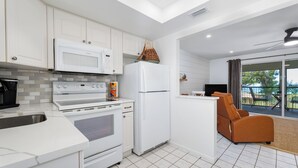 Cocina moderna con armarios blancos, microondas, estufa y refrigerador. Cuenta con una zona de estar con una silla marrón y vistas al exterior a través de una puerta de vidrio.