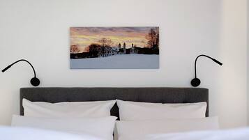 Ein Bett mit weißer Bettwäsche, einem grauen Kopfteil und einem Wandbild, das einen Sonnenuntergang über einem Gebäude zeigt.