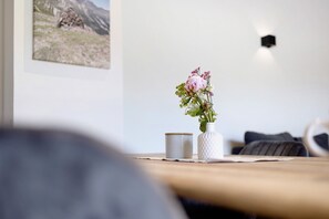 A room with a wooden table, a white vase with flowers, a grey mug, and a framed picture on the wall. - Apartment 'wie Dahoam' – 'Schwaiger Almblick' with Mountain View, Private Terrace & Wi-Fi (Bad Tölz)