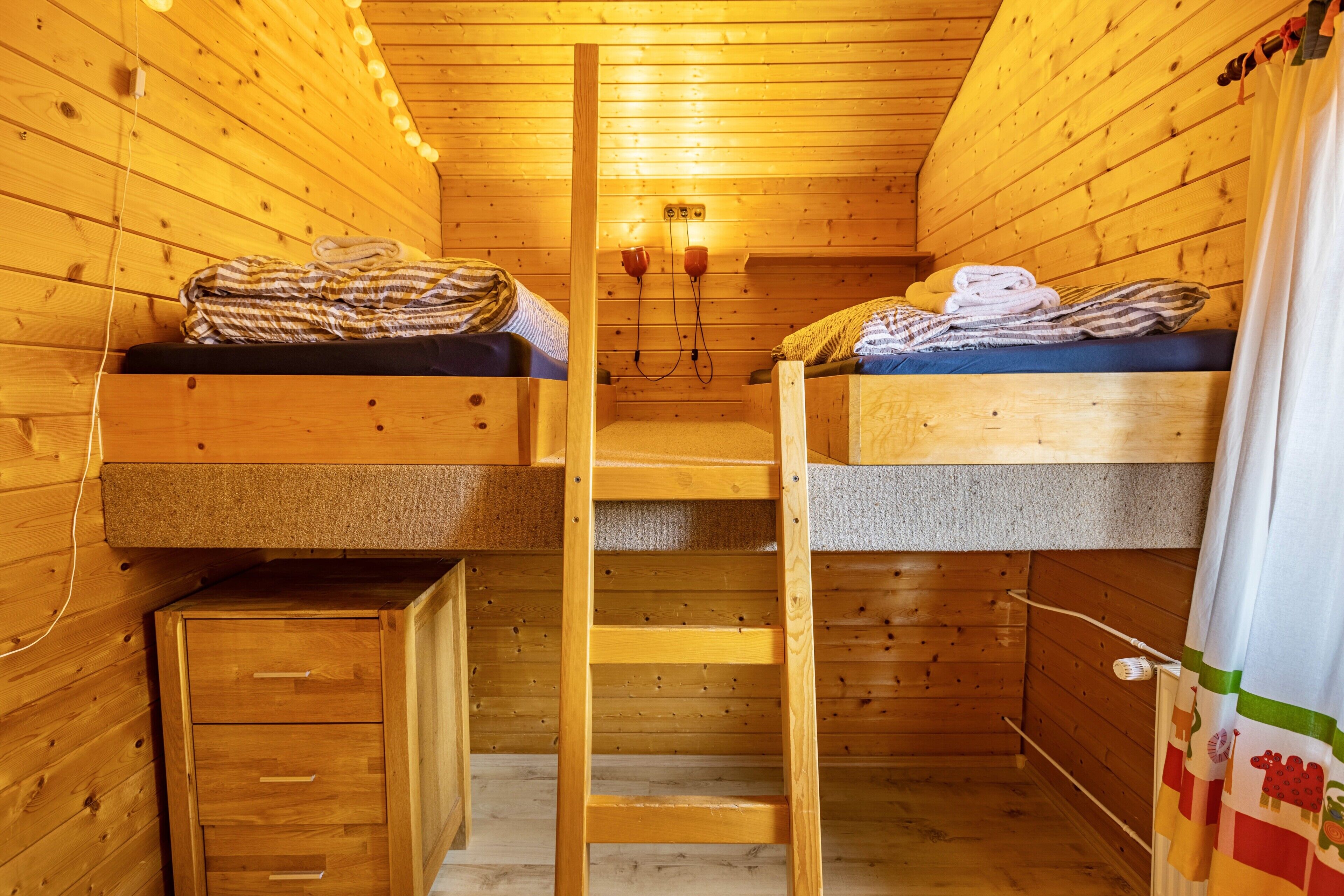 Une pièce de cabane en bois avec des lits superposés, une échelle, un petit placard et un luminaire fixé au mur.