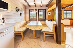 Un intérieur en bois comprenant une table à manger, des bancs, un coin cuisine et de grandes fenêtres.