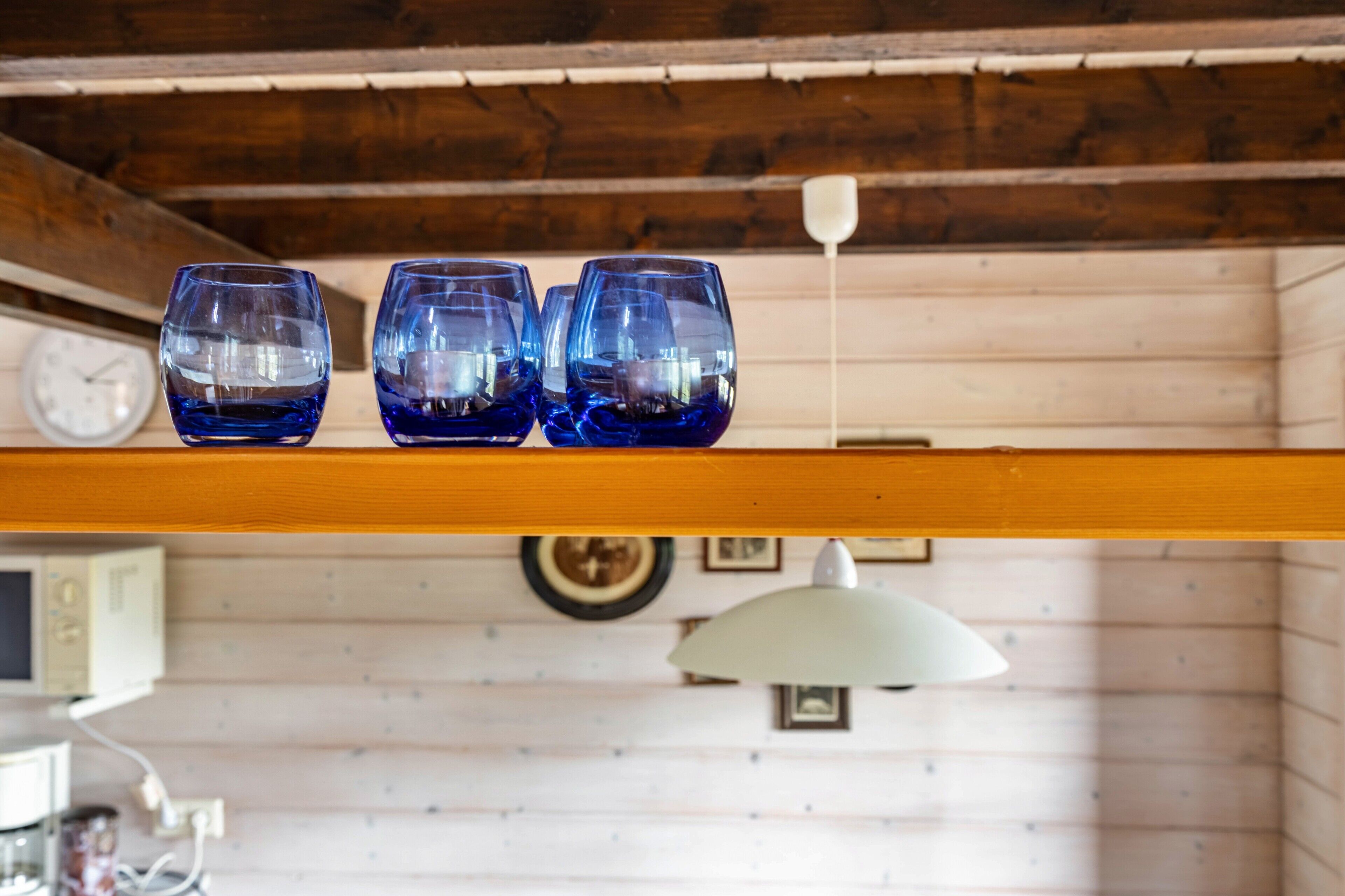 Three blue glasses on a wooden shelf in a room with wooden walls, a clock, and a hanging lamp.