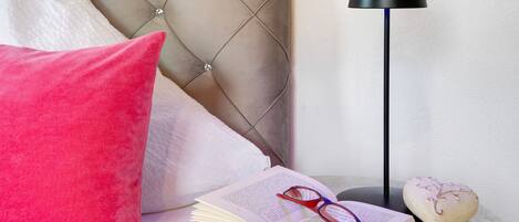 A bed with a tufted headboard, a red pillow, a bedside table with a book, glasses, and a decorative object.