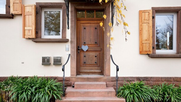 A wooden front door with a heart-shaped decoration, a set of steps leading up to it, and two potted plants on either side.