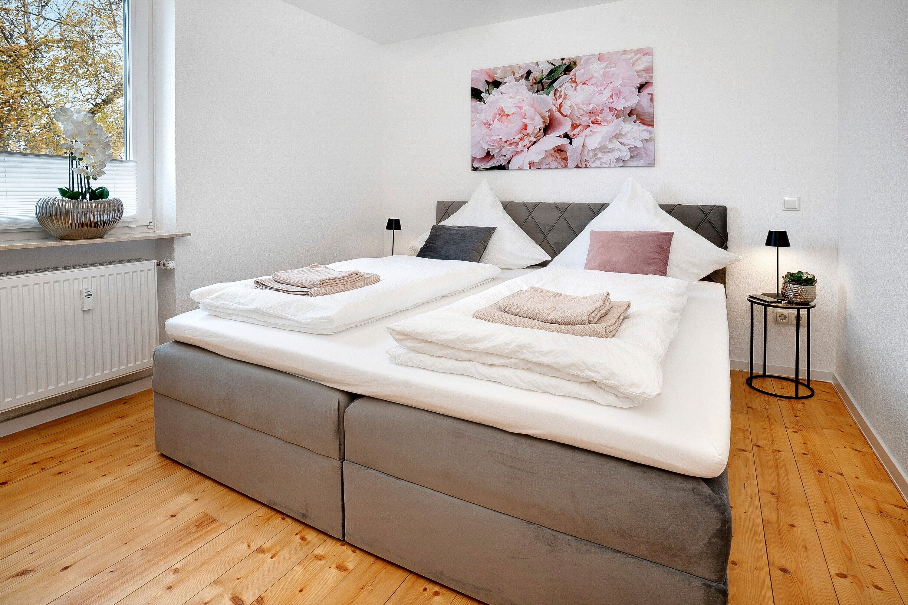 Un letto matrimoniale con testiera grigia rivestita in tessuto, biancheria da letto bianca e un grande quadro con fiori sopra.