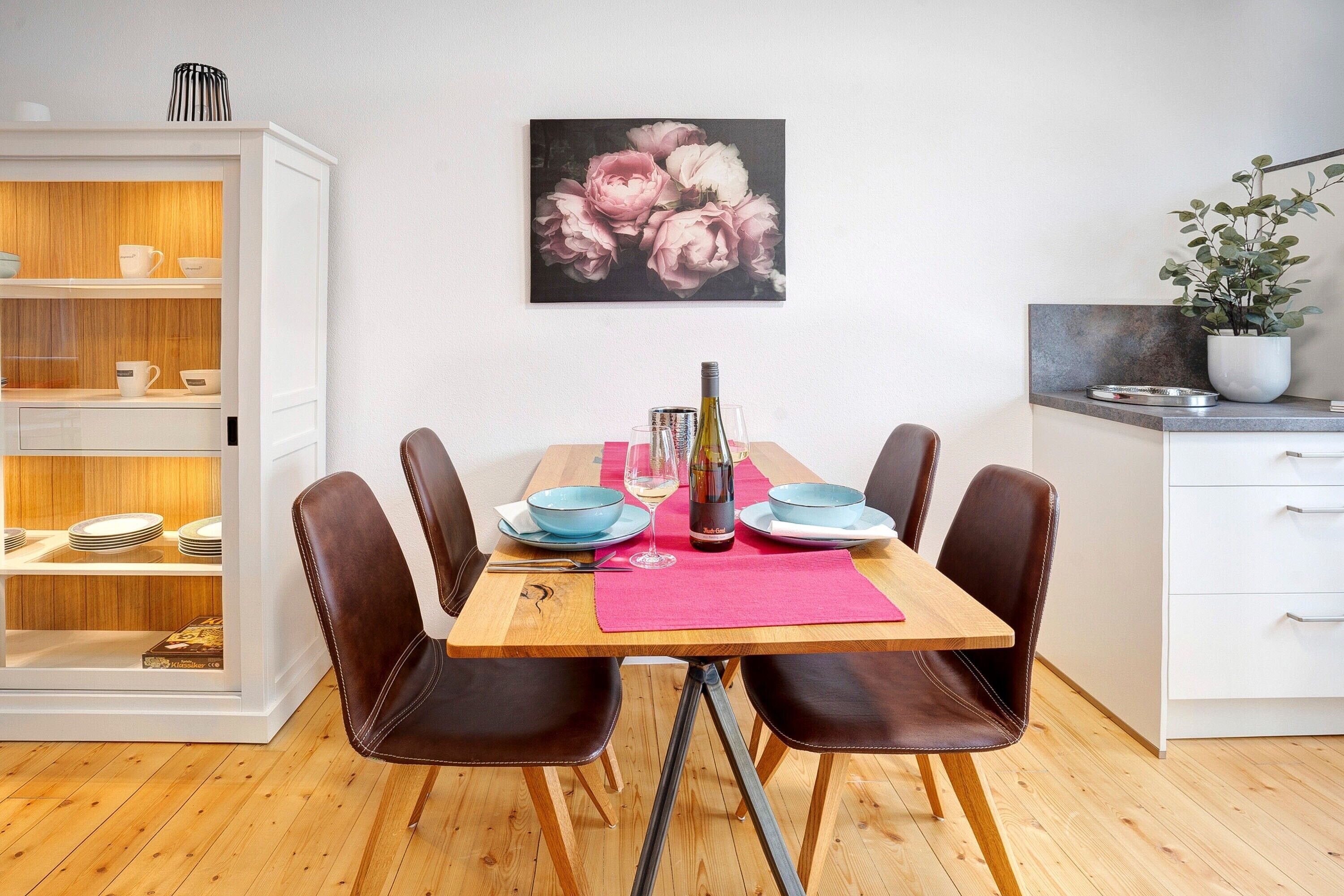 A dining area with a wooden table, four chairs, a pink placemat, and a painting of flowers on the wall.