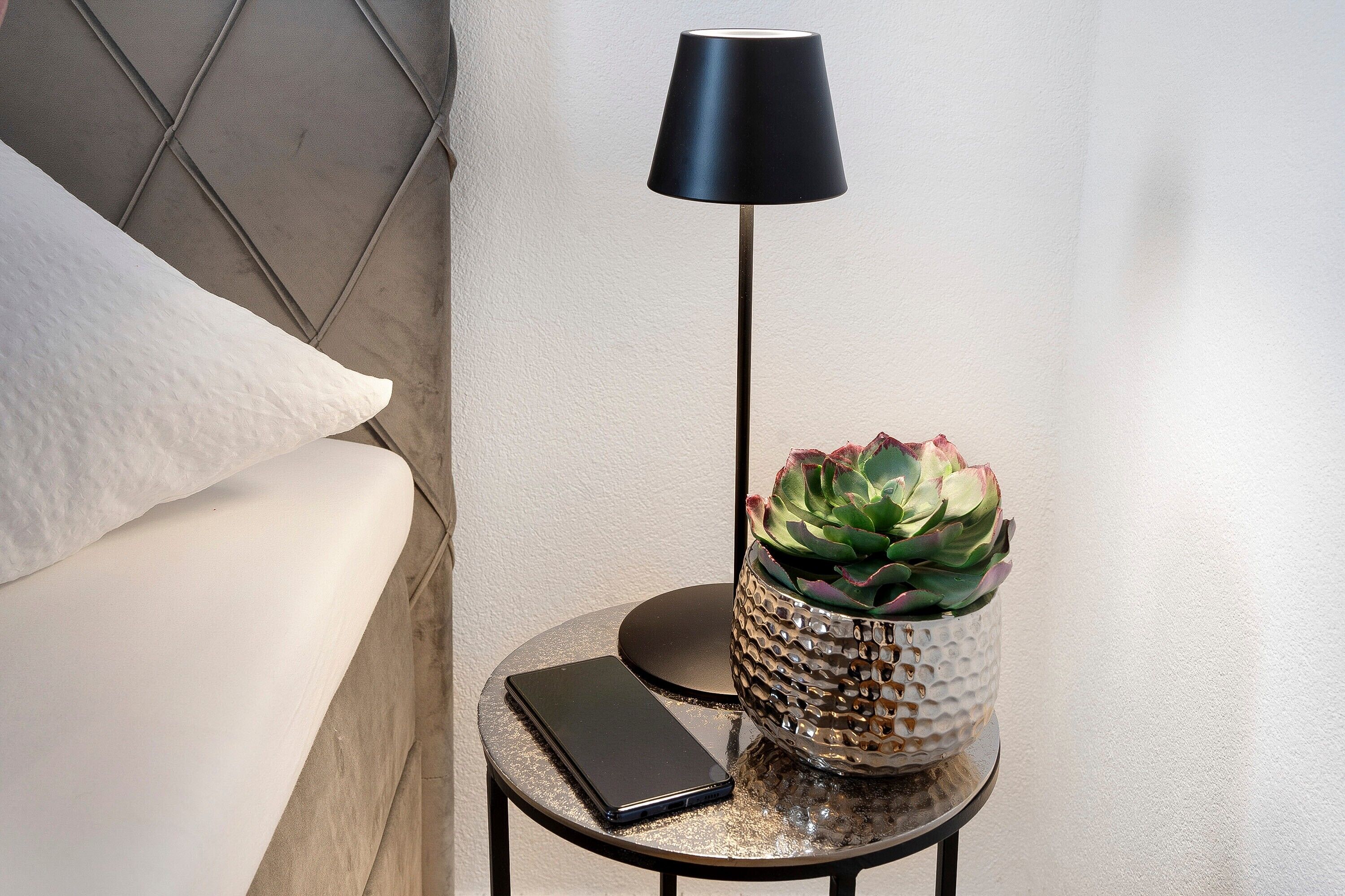 Una cama con un almohadón, una mesita redonda con una lámpara, un teléfono y una planta decorativa.