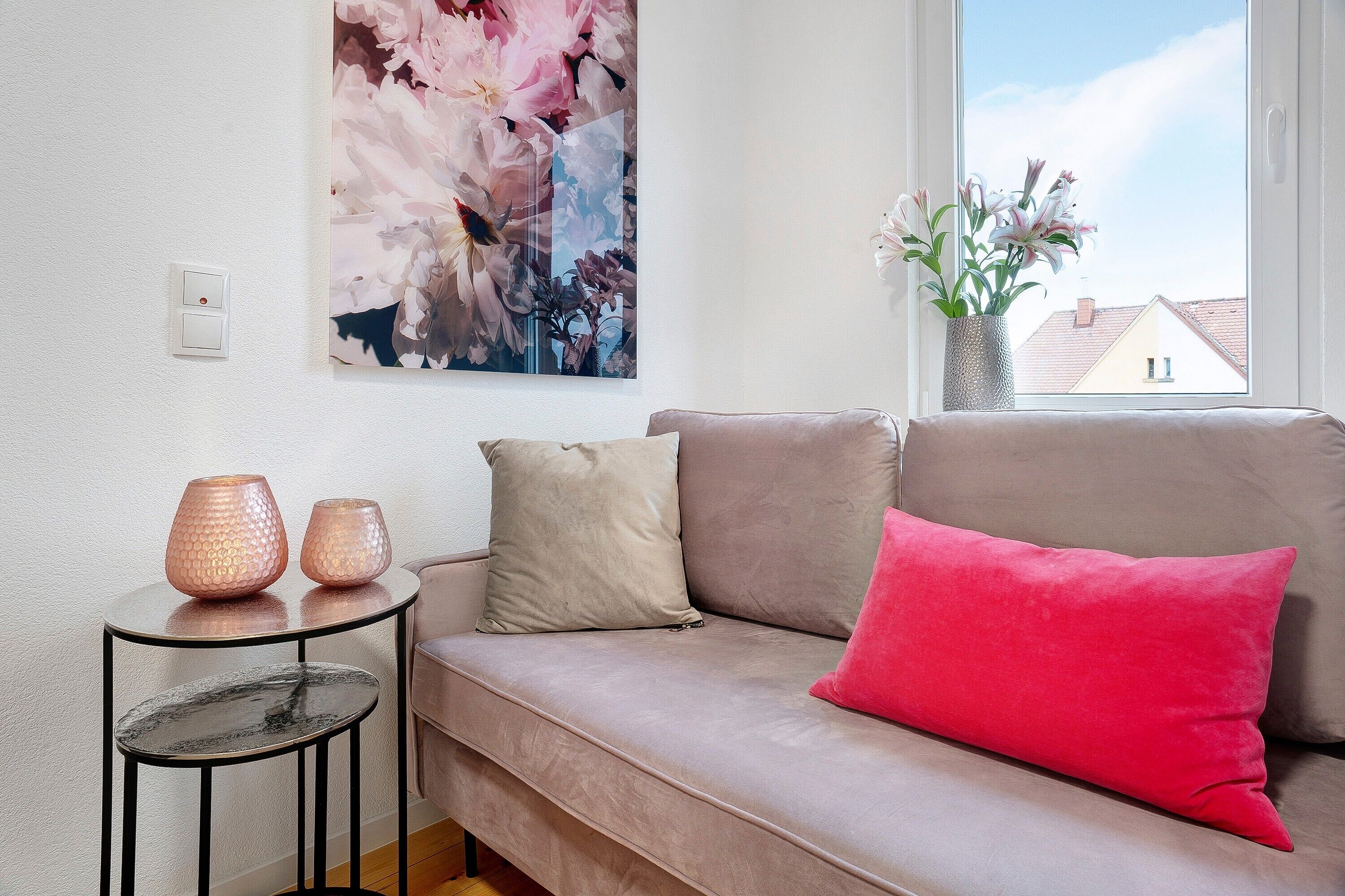 Una sala de estar con un sofá, una mesita con dos velas, un jarrón con flores y un cuadro grande con motivos florales en la pared.