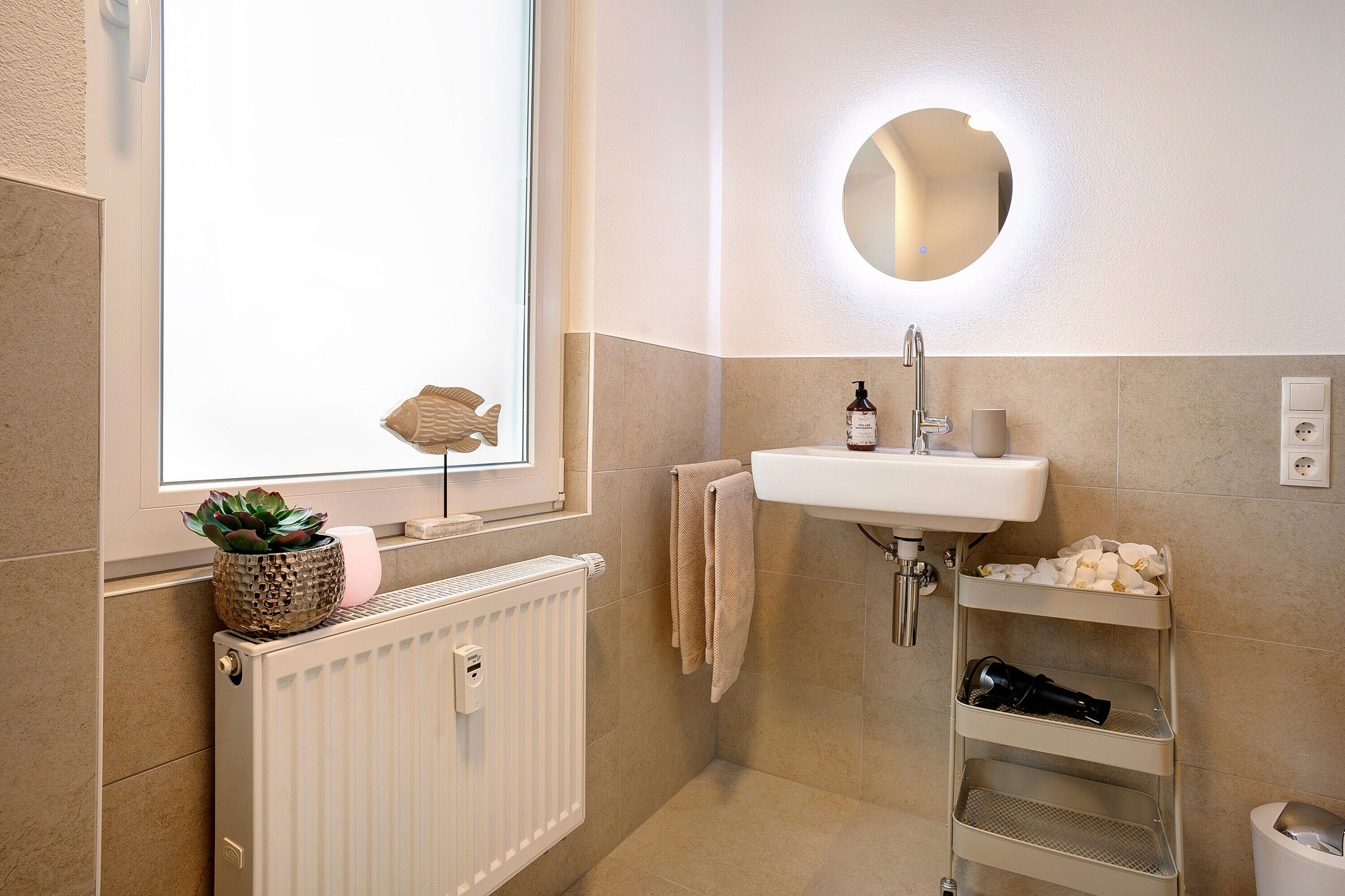 Une salle de bain avec une fenêtre, un lavabo, un miroir, un radiateur et une étagère contenant des articles de toilette.