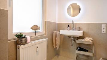 Une salle de bain avec une fenêtre, un lavabo, un miroir, un radiateur et une étagère contenant des articles de toilette.