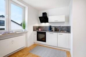 Eine kleine Küche mit weißen Schränken, einem eingebauten Backofen, einer schwarzen Dunstabzugshaube, einem Fenster und einem gemusterten Teppich.