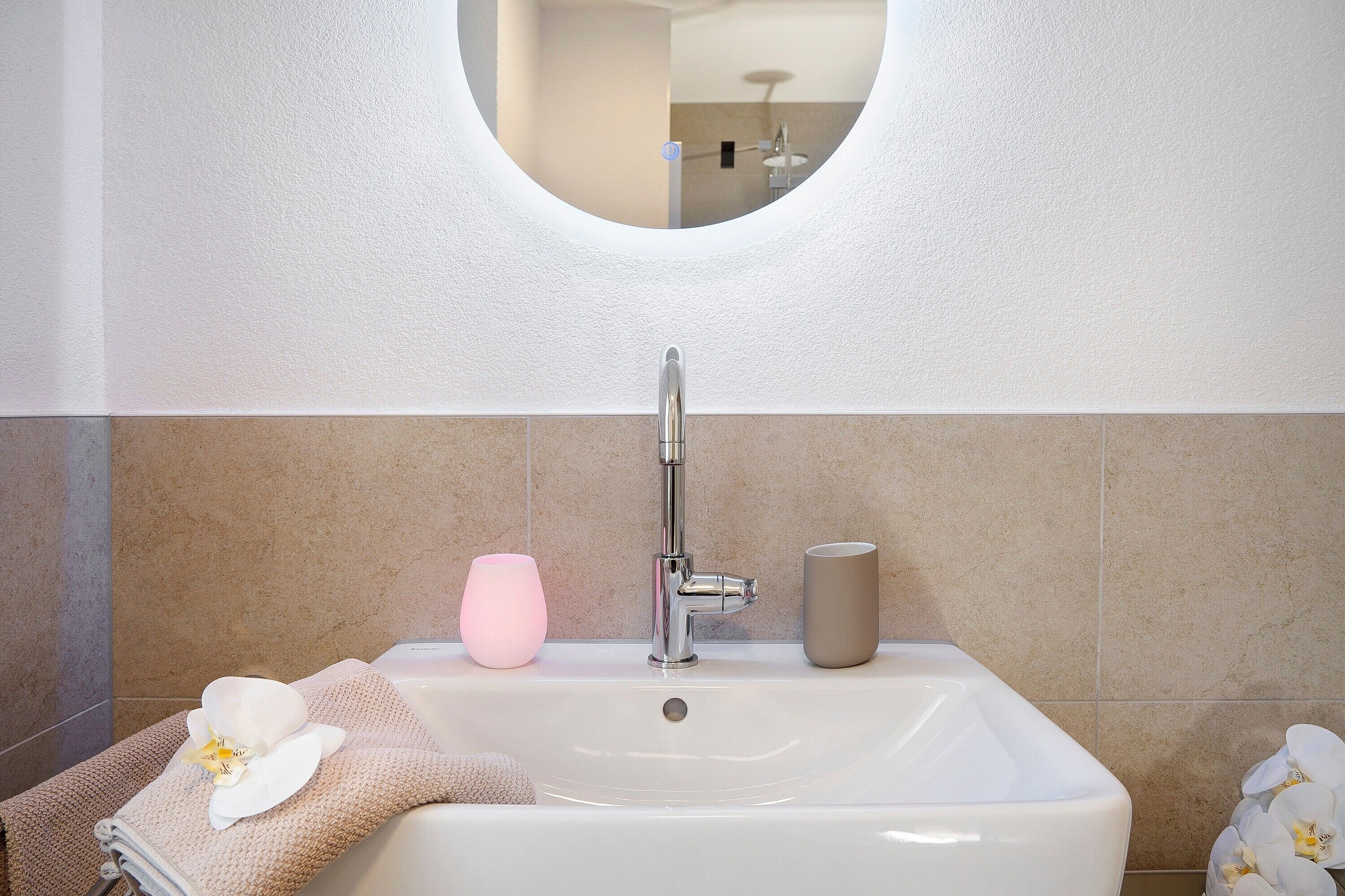 A bathroom with a white sink, a round mirror, and a towel with an orchid.