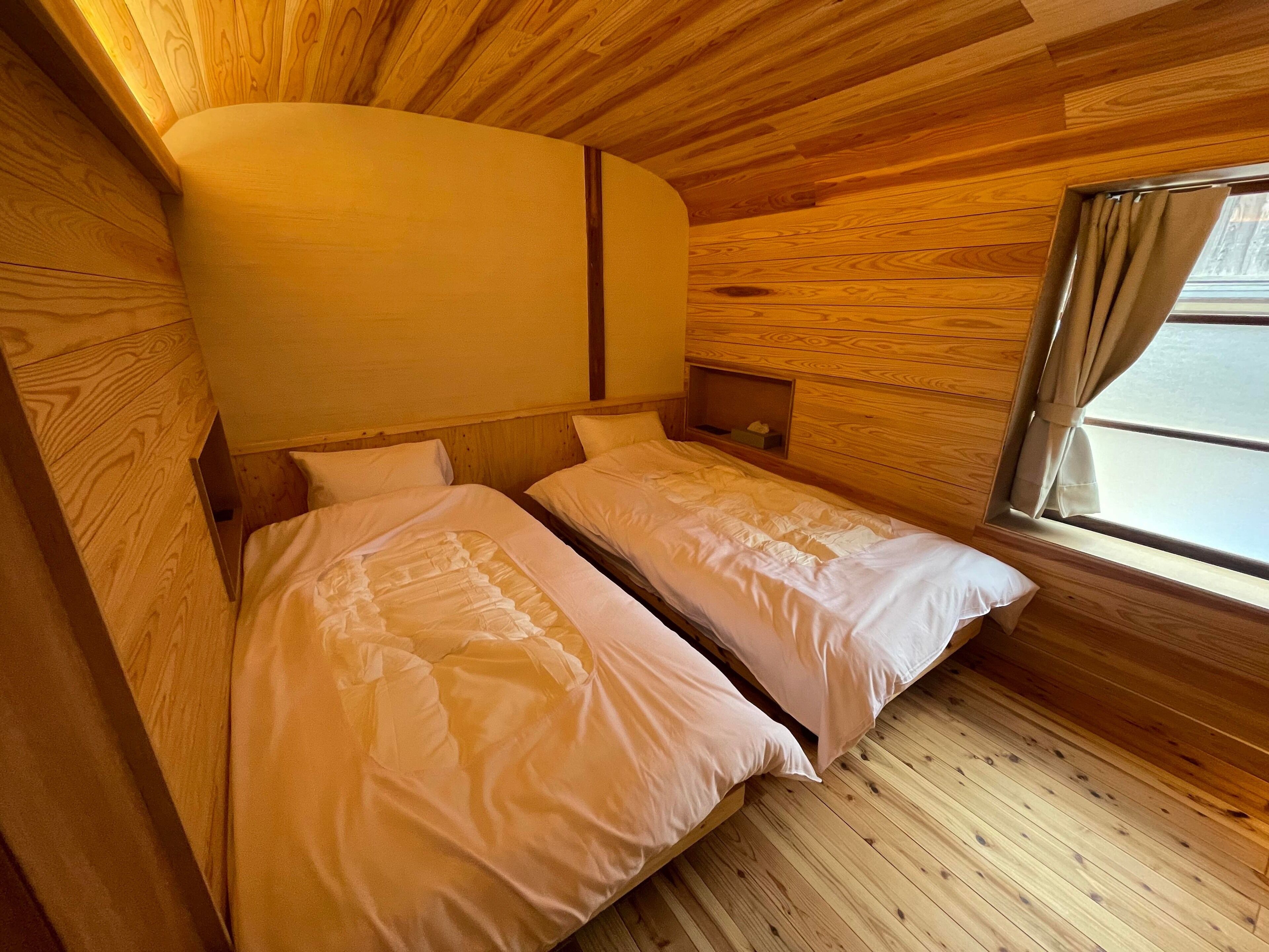 A small, cozy room with two beds, wooden walls, and a window with curtains.