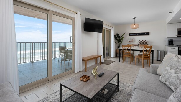 Un salon moderne doté d’une grande porte-fenêtre coulissante donnant sur un balcon, d’un coin repas avec des meubles en bois et d’une cuisine avec des meubles blancs.