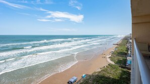 On peut admirer la plage depuis un balcon donnant sur une étendue de sable bordée de vagues, quelques parasols et un ciel dégagé.