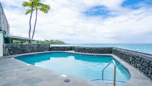 A swimming pool with a stone wall and a metal handrail, surrounded by a stone deck and palm trees, with a view of the ocean. - spectacular ocean front studio condo,  Magic sands, ideal view and location (Kailua-Kona)