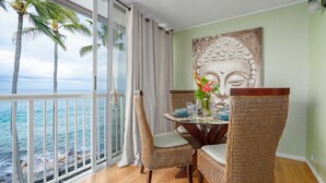 A balcony with a view of the ocean, wicker chairs, a table set for dining, and a Buddha statue on the wall. - spectacular ocean front studio condo,  Magic sands, ideal view and location (Kailua-Kona)