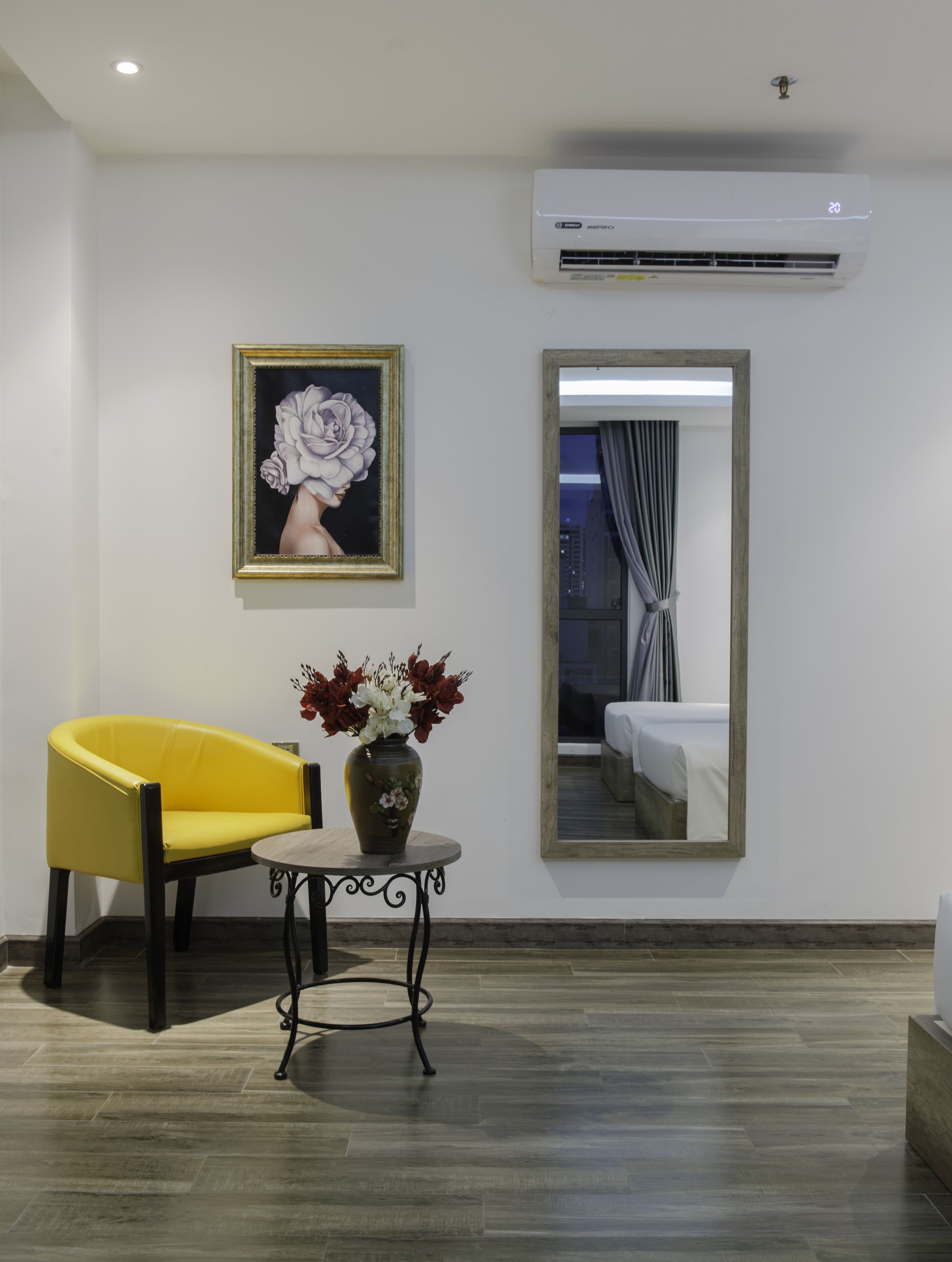 Une pièce avec une chaise jaune, un vase de fleurs, un tableau encadré et un grand miroir qui reflète un lit et des rideaux.