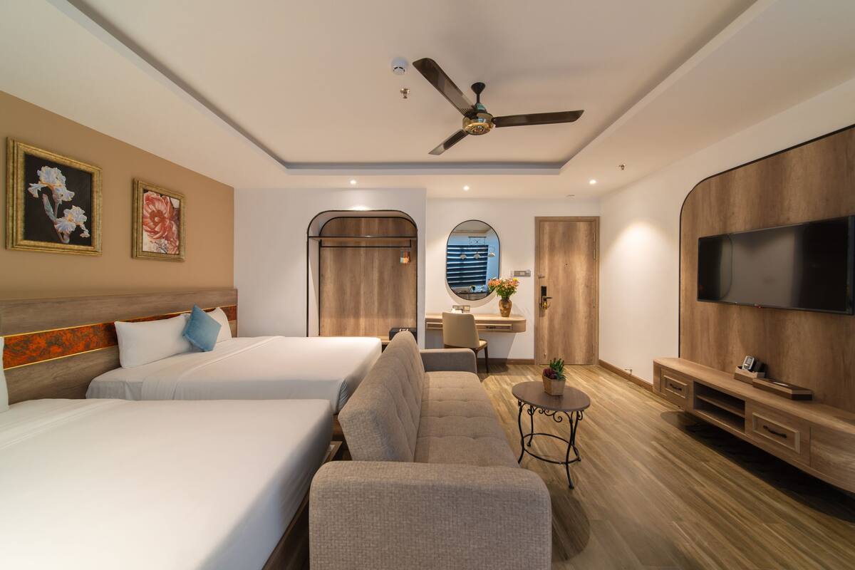 A hotel room with two beds, a sofa, a TV, a ceiling fan, and a small table with a plant.