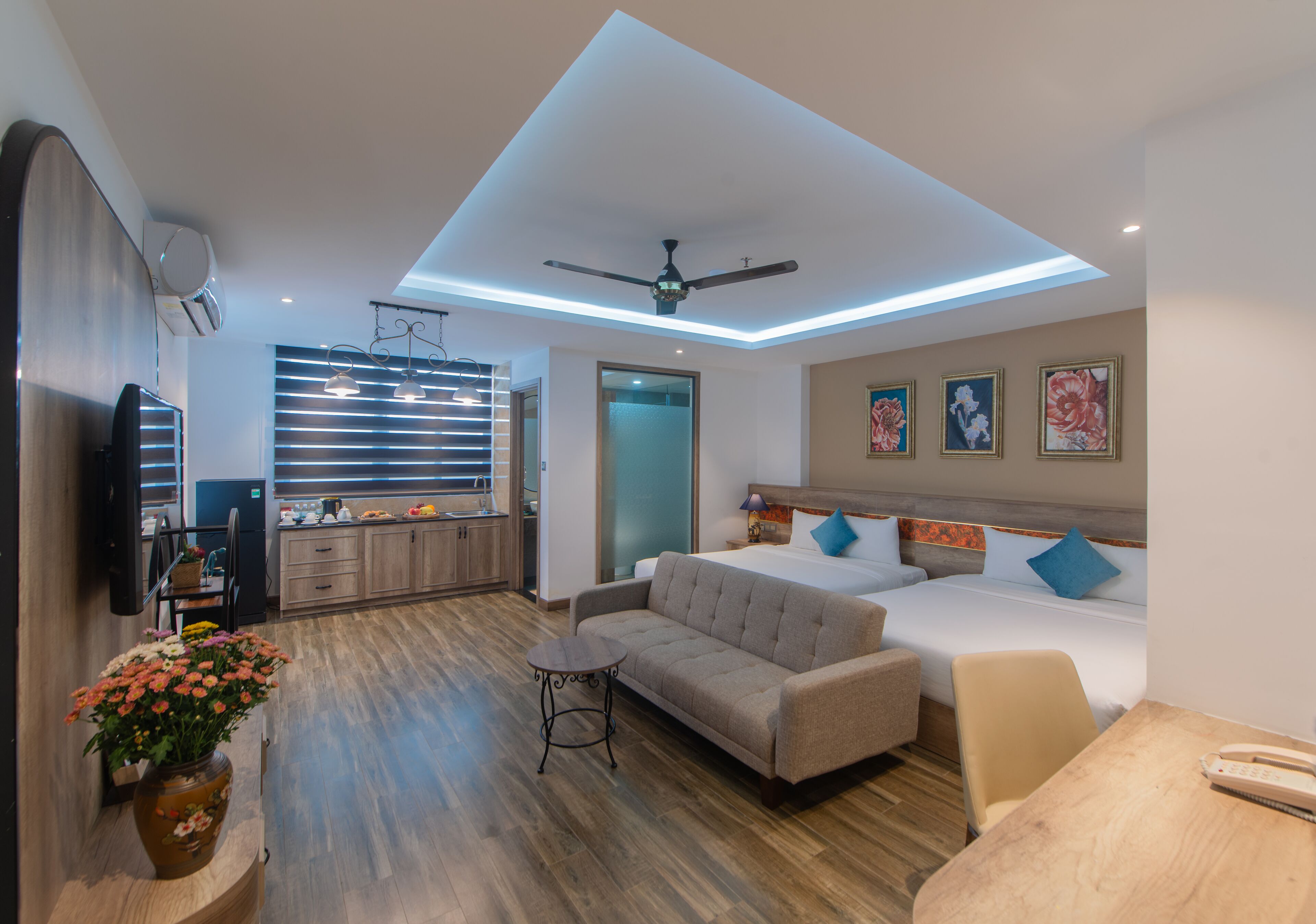 A modern hotel room with a sofa, a small table, a bed with blue pillows, a ceiling fan, and a kitchen area in the background.