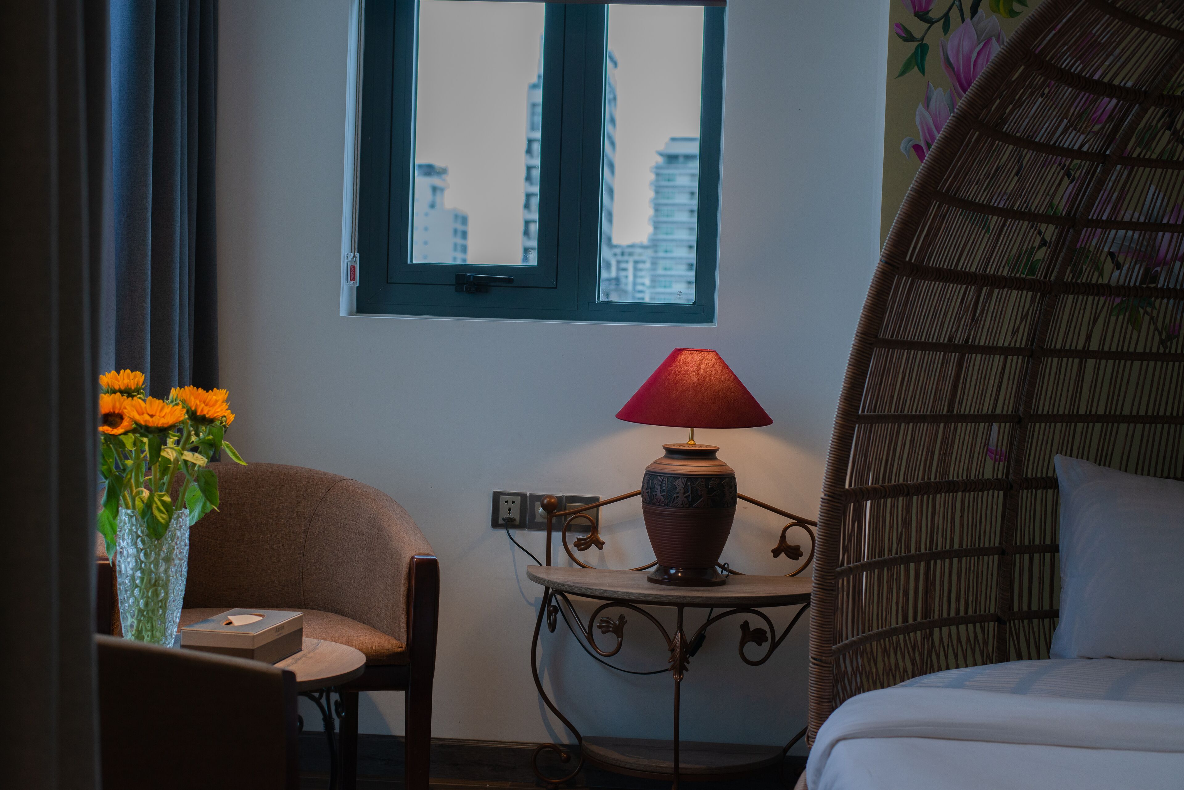 A room with a bed, a chair, a table with a lamp, a vase with flowers, and a window with a view of buildings.