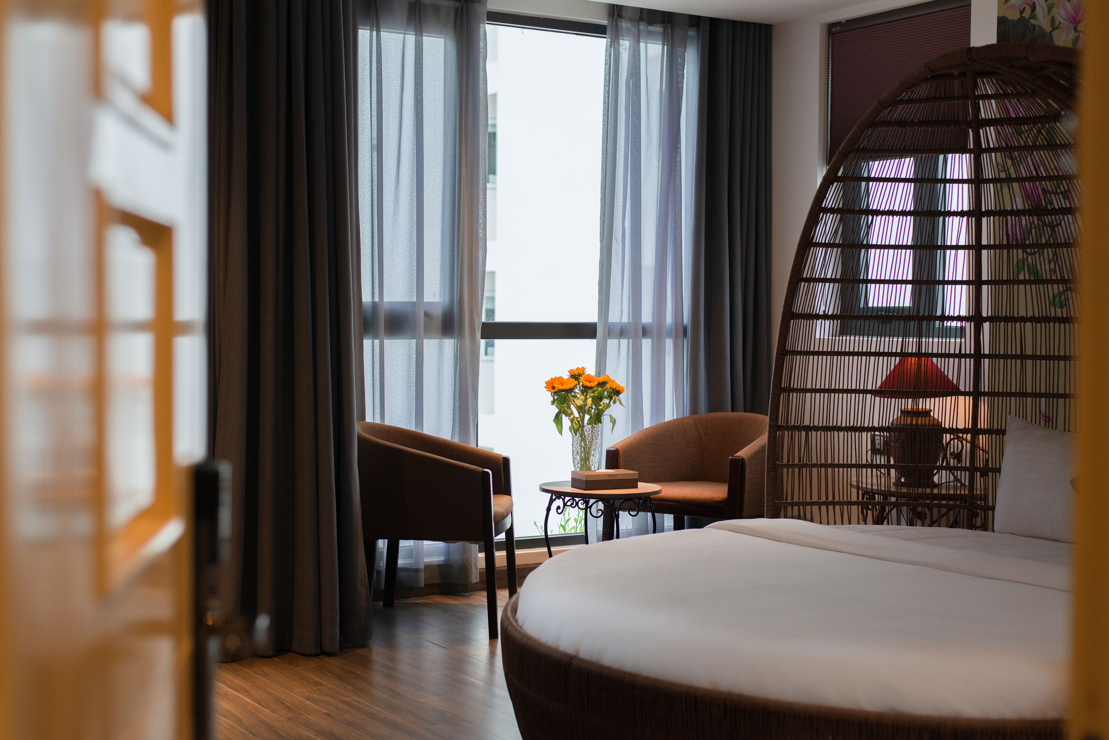 A hotel room with a large bed, two chairs, a small table with a vase of flowers, and a view through sheer curtains to the outside.