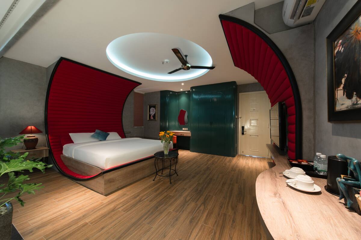 A modern hotel room with a curved bed, a ceiling fan, a small table with a vase, and a curved wooden counter with cups.
