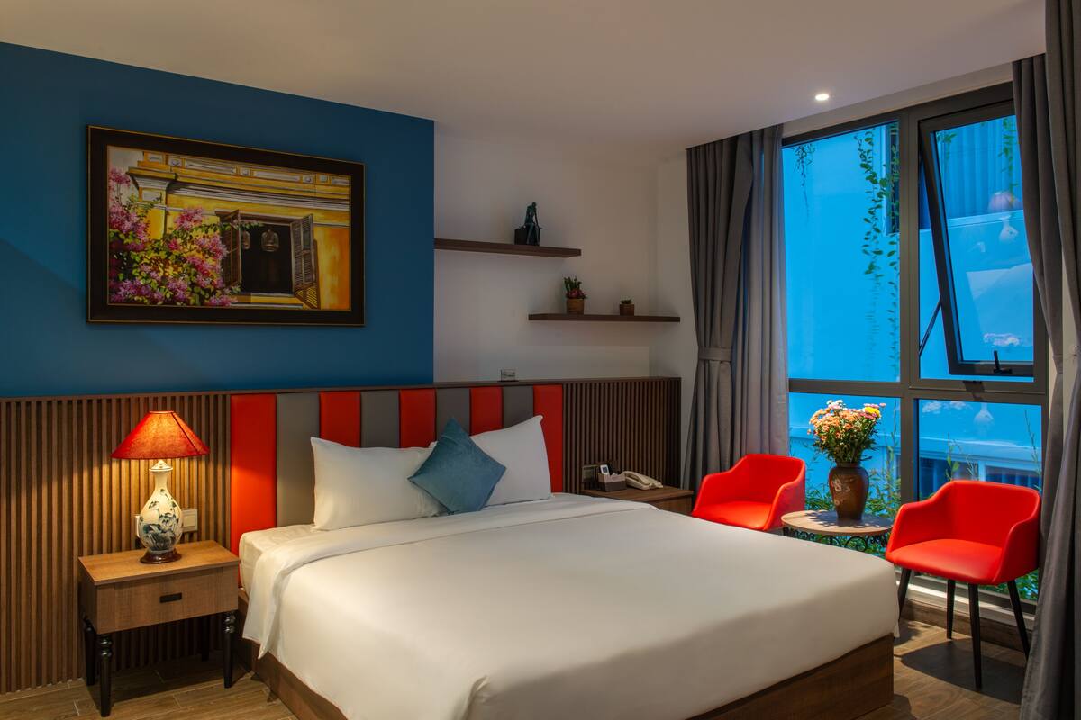 A hotel room with a bed, a nightstand with a lamp, two red chairs, a small table with a vase, and a window with curtains.