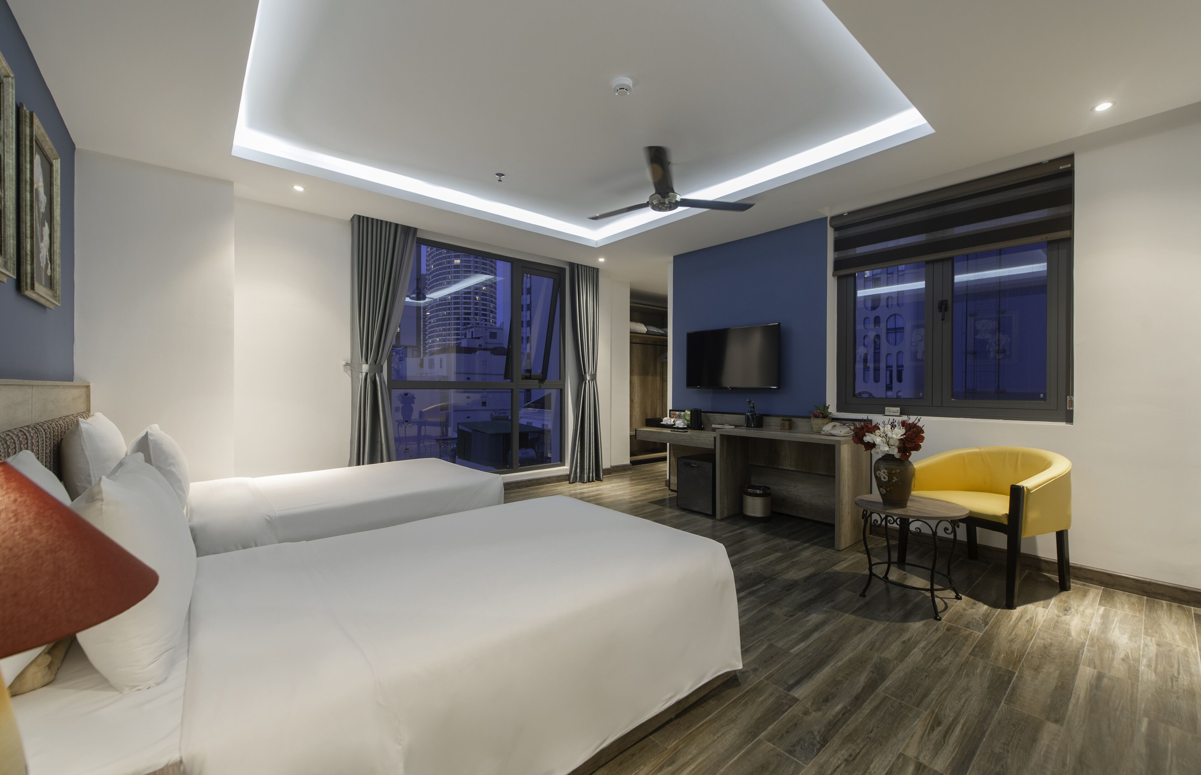 Quarto de hotel moderno com uma cama grande, uma escrivaninha, uma cadeira, uma televisão e uma janela com vista para a cidade.