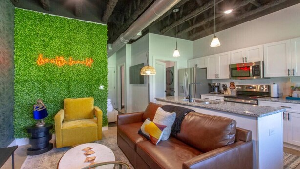 Una sala de estar moderna con una pared verde, un sillón amarillo, un sofá de cuero marrón y una cocina con armarios blancos y electrodomésticos de acero inoxidable.