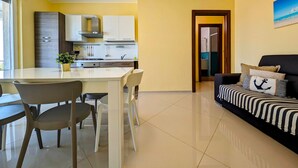 Eine moderne Küche mit weißen Schränken und einer zentralen Kochinsel, ein Essbereich mit Stühlen und ein Wohnzimmer mit einer Couch und gestreiften Kissen.