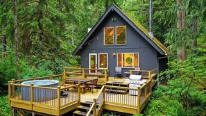 A cabin with a deck, hot tub, and outdoor seating area surrounded by forest.