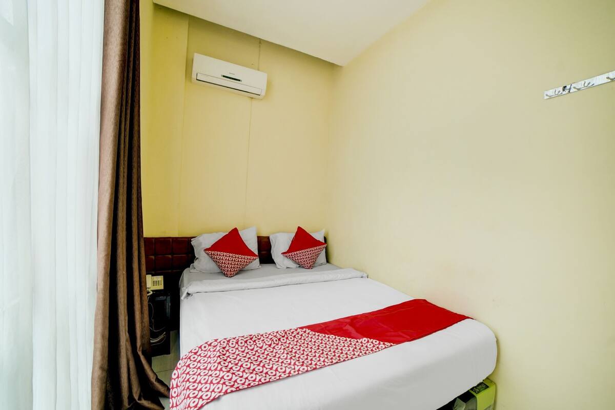 Una camera d'albergo con un letto, una coperta rossa e bianca a motivi geometrici, due cuscini rossi, un condizionatore d'aria bianco e un telefono sul comodino.
