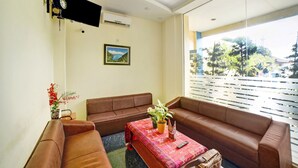 A living room with a brown sofa set, a small table with a plant, a painting on the wall, and a large window with a view of trees and buildings. - Hotel O Wesly House (Medan)
