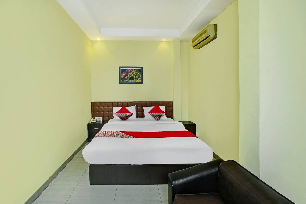 Una camera d'albergo con un letto grande, una testiera, un comodino, una sedia e un condizionatore d'aria fissato alla parete.