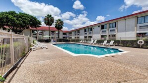 A swimming pool with lounge chairs, a fenced area, and a building with multiple windows and a red roof.