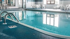 A swimming pool with clear blue water, surrounded by white chairs and tables, and a tiled wall with a window. - Baymont by Wyndham Flint Airport North (Flint)