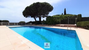 Ein rechteckiger Swimmingpool mit klarem, blauem Wasser, umgeben von einer gefliesten Terrasse. Im Hintergrund sind Bäume und ein Zaun zu sehen.