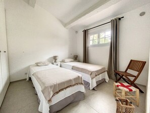A small, single-room accommodation with two single beds, a wooden chair, and a window with curtains.