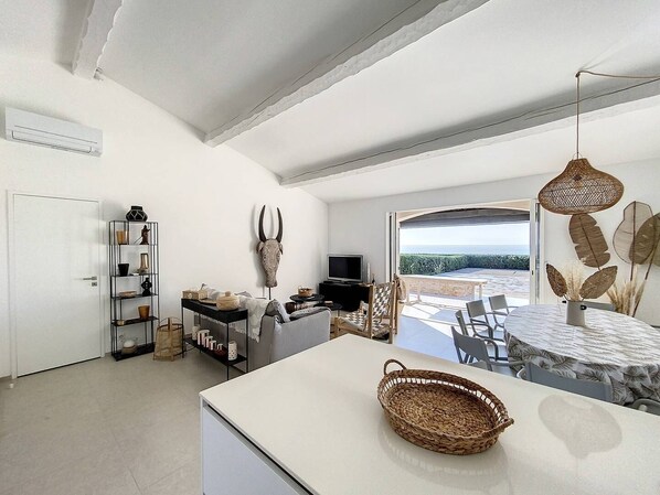 A modern living room with a white sofa, a TV, and a dining area with a table and chairs. There is a large window with a view of the ocean.
